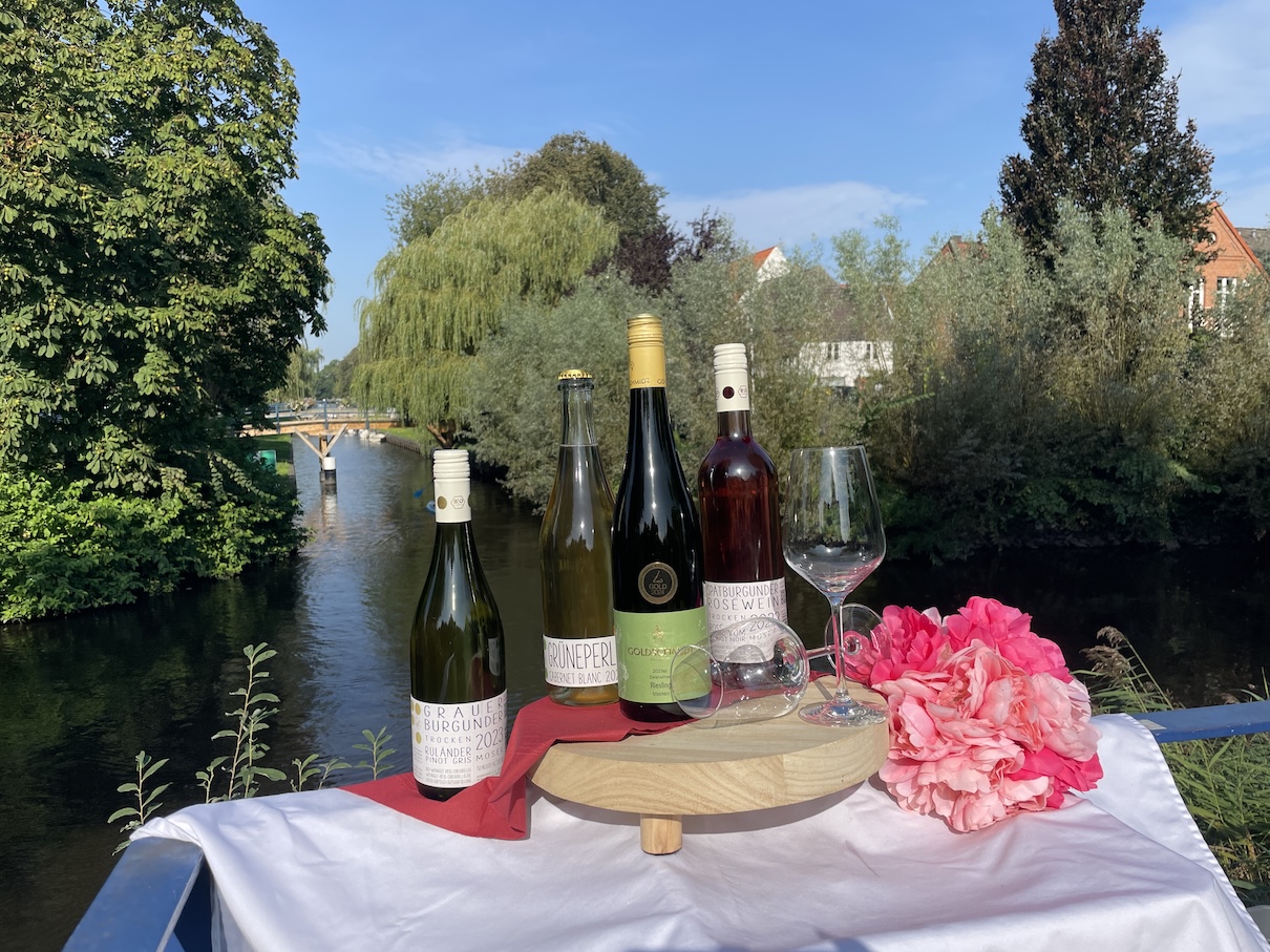 Verschiedene Weinflaschen und zwei Weingläser stehen auf einem Holzbrett auf einer Brücke.